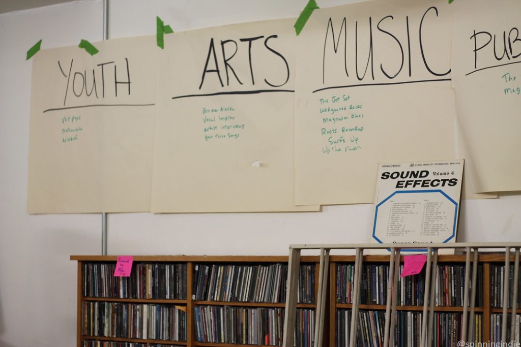 CD shelves full of CDs, with Sound Effects LP atop the shelf. Paper signs above read: "youth", "arts," "music" and "pub..." with lists of programs written beneath each category. At Space 101.1 FM. Photo: J. Waits/Radio Survivor