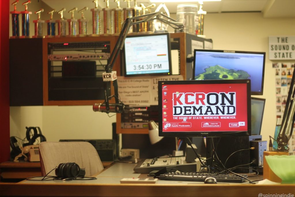 Photo of studio of college radio station KCR. Pictured: computer monitor that reads "KCR On Demand. The Sound of State. Wherever. Whenever," sound board, computer keyboard, headphones, and microphone. Overhead is a shelf lined with trophies in the shape of microphones. Photo: J. Waits/Radio Survivor