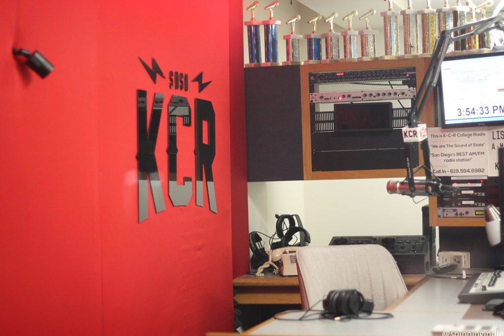 Photo of studio at college radio station KCR. On the left is a red wall with a SDSU KCR sign. A desk and chair are positioned near a hanging microphone, with computer monitor and other audio equipment above and a shelf of microphone-shaped trophies above that. Photo: J. Waits/Radio Survivor