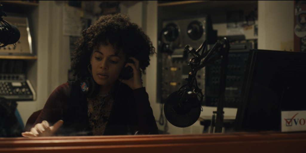 Screen grab from Mare of Easttown. Kiah McKirnan (as DJ Anne Harris) wears headphones in booth of Haverford College radio station WWXU. There's a microphone in front of her and a reel-to-reel machine behind her. 