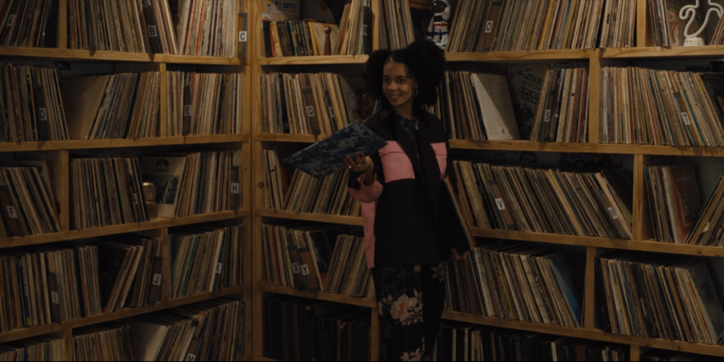 Screen shot of Mare of Easttown record library at Haverford College radio station. Kiah McKirnan (as Anne Harris) holds an LP in her hand while standing in front of huge shelves full of vinyl records.