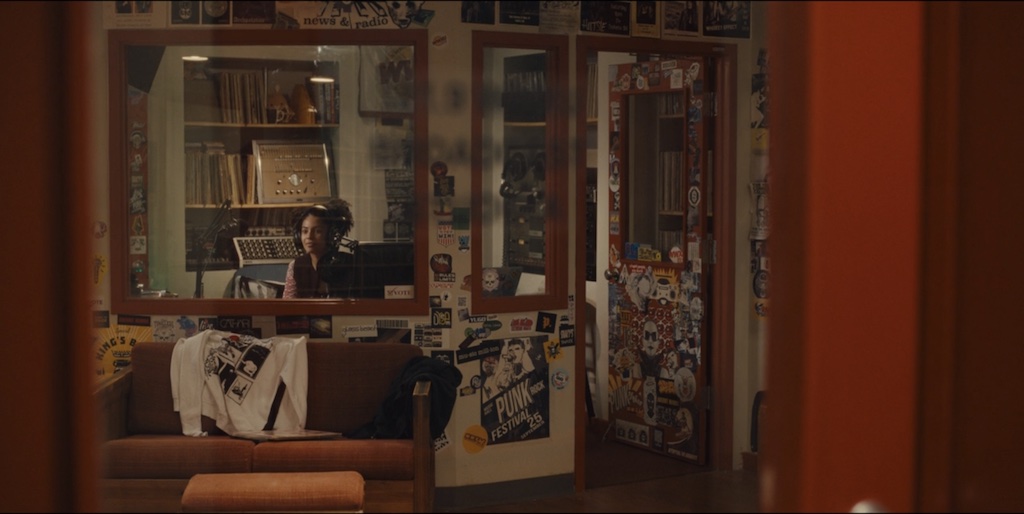 Screen grab from Mare of Easttown. Kiah McKirnan (as Anne Harris) sits in the WWXU booth. The viewer looks in through the window of the station door and we can see a T-shirt flung over an orange couch, sticker and poster-covered walls and door, and audio equipment behind the DJ in the booth.