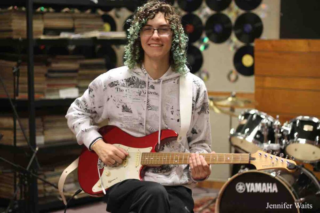 Person wearing glasses with curly hair, dyed green on the ends. They are playing a red and white guitar and there's a drum set to their left. Vinyl records are decorated the wall behind them. Photo: J. Waits