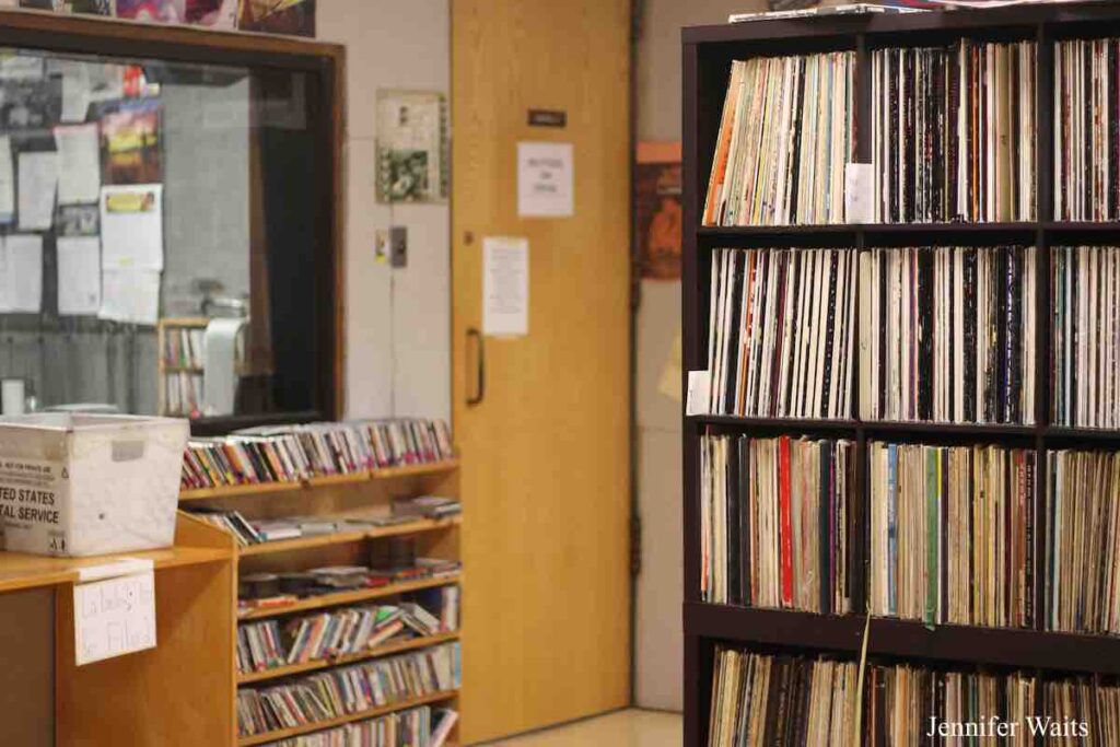 Room at college radio station WRPI. On the right are black shelves full of vinyl record albums. To the left is a window with a view to another studio. Below that are shelves with CDs on them. A US mail bin is to the left of that. Photo: J. Waits