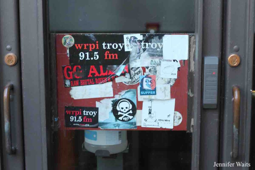 Photo of exterior door with sticker covered panel that has WRPI Tory 91.5 FM sticker, a WRPI sticker with a skull on it and layers of other stickers and peeling off stickers. Photo: J. Waits