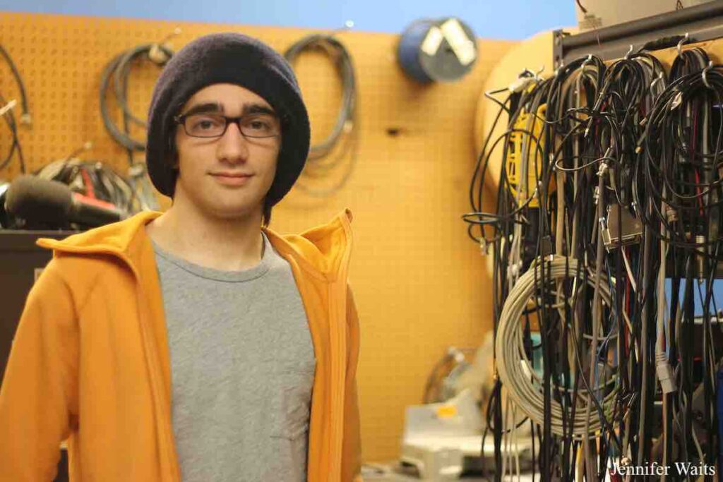 Man wearing black beanie cap and glasses, with a grey Tshirt and orange sweatshirt. He's standing in front of a peg board with cabling attached to it. Next to him is a wall full of looped cables. Photo: J. Waits