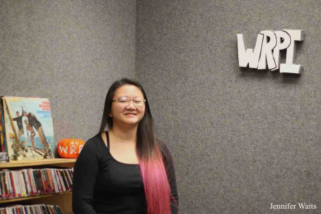 Woman with long black and dyed pink hair, wearing glasses and black clothing. Behind her is a shelf with CDs, a pumpkin, and a vinyl record. A carpeting grey wall behind her has a 3-D white and grey sign with letters WRPI. Photo: J. Waits