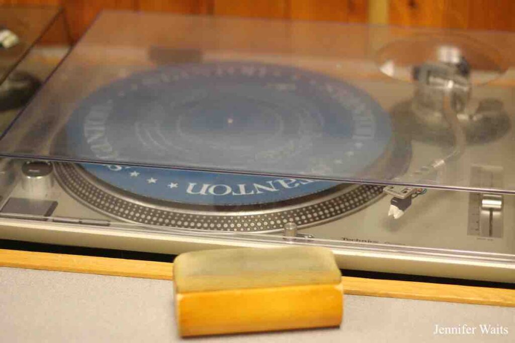 Photo of turntable with plastic case covering it. In front of turntable there's a wood and fabric record cleaning brush. Photo: J. Waits