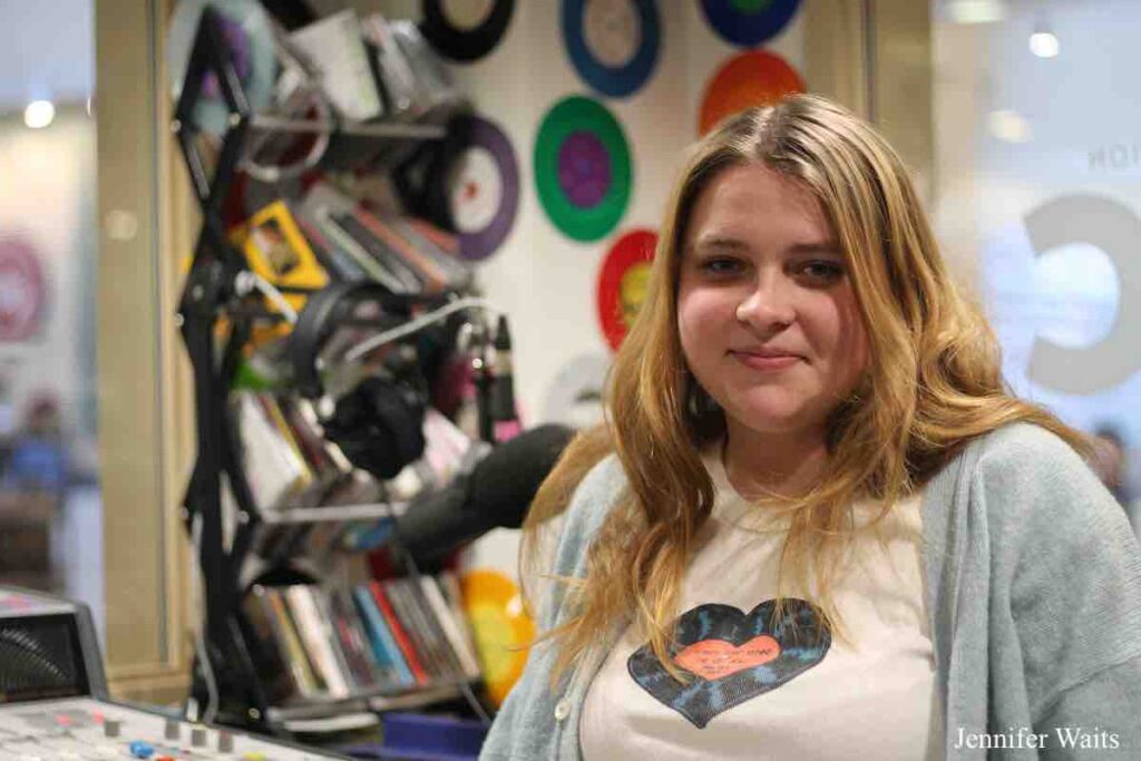 Woman with long blonde hair, wearing blue cardigan and white T-shirt with a heart shaped record drawing on it. Behind here there are colorful vinyl records on the wall, a small set of shelves with CDs, and a pair of headphones. Photo: J. Waits