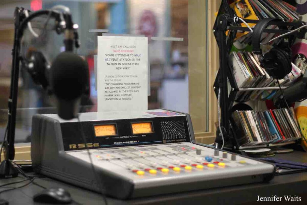 Photo of WRUC's studio in Student Center. Small mixing board at center. Microphone to left. Rack on CDs on the right, with a pair of headphones hanging nearby. Photo: J. Waits