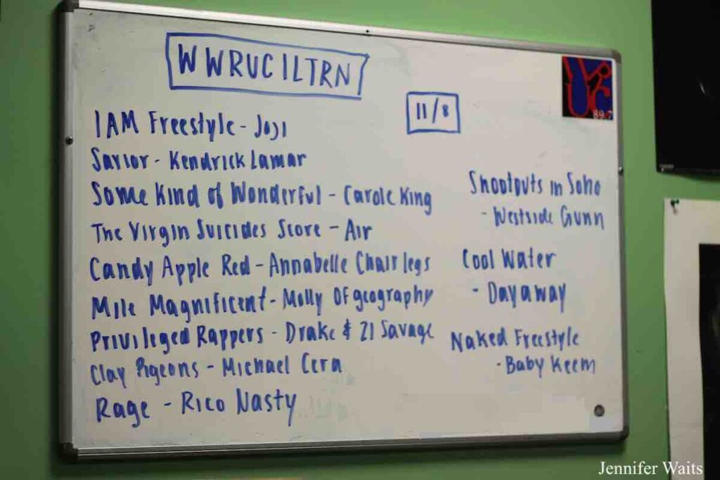 Photo of white dry erase board hanging on a green wall. Board reads: "WWRUC ILTRN 11/8" and then lists song titles and artists. A few of the artists include Kendrick Lamar, Carole King, Michael Cera, and Rico Nasty. Photo: J. Waits