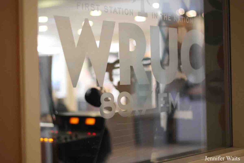 Photo of the outside of college radio station WRUC's downstairs studio. Image is looking into a window. WRUC 89.7 is written on the window and one can see lights from audio equipment in the room. Photo: J. Waits