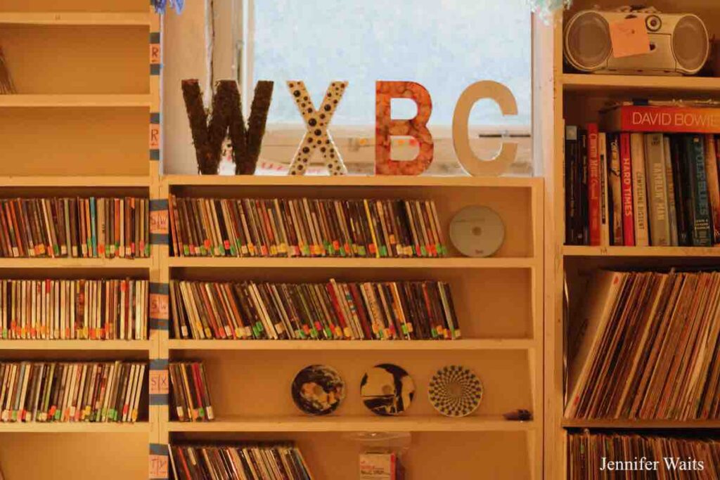 Photo of shelves at college radio station WXBC. On the shelves are many CDs in cases and some outside of cases. Decorated call letters WXBC near a window. Books and LPs on shelves to the right. Photo: J. Waits