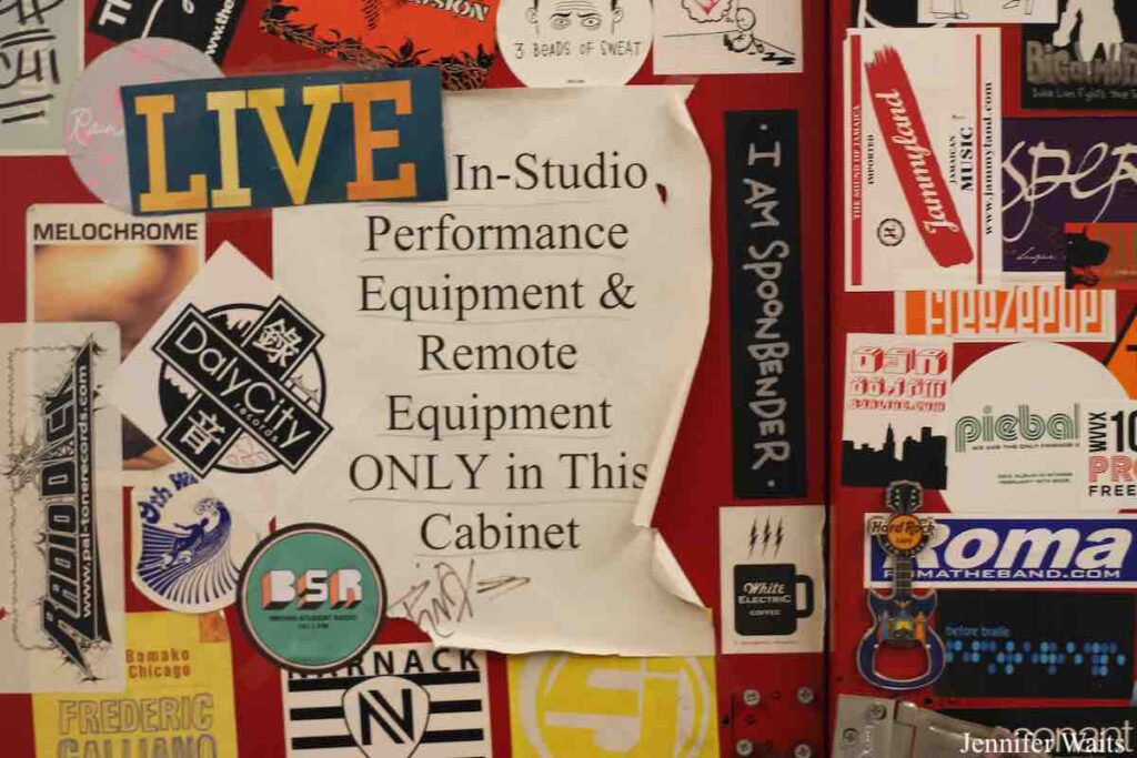 Photo of Cabinet at college radio station BSR. It has a sign that reads "In-Studio Performance Equipment & Remote Equipment ONLY in this Cabinet. Stickers for BSR and for bands surround that sign. Photo: J. Waits