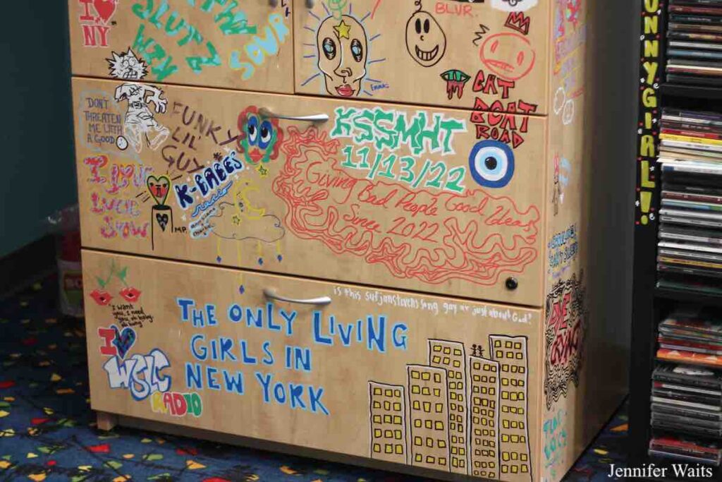 Photo of college radio station WSLC. Paintings and drawings on wooden cabinet with black shelf of CDs next to it. Photo: J. Waits