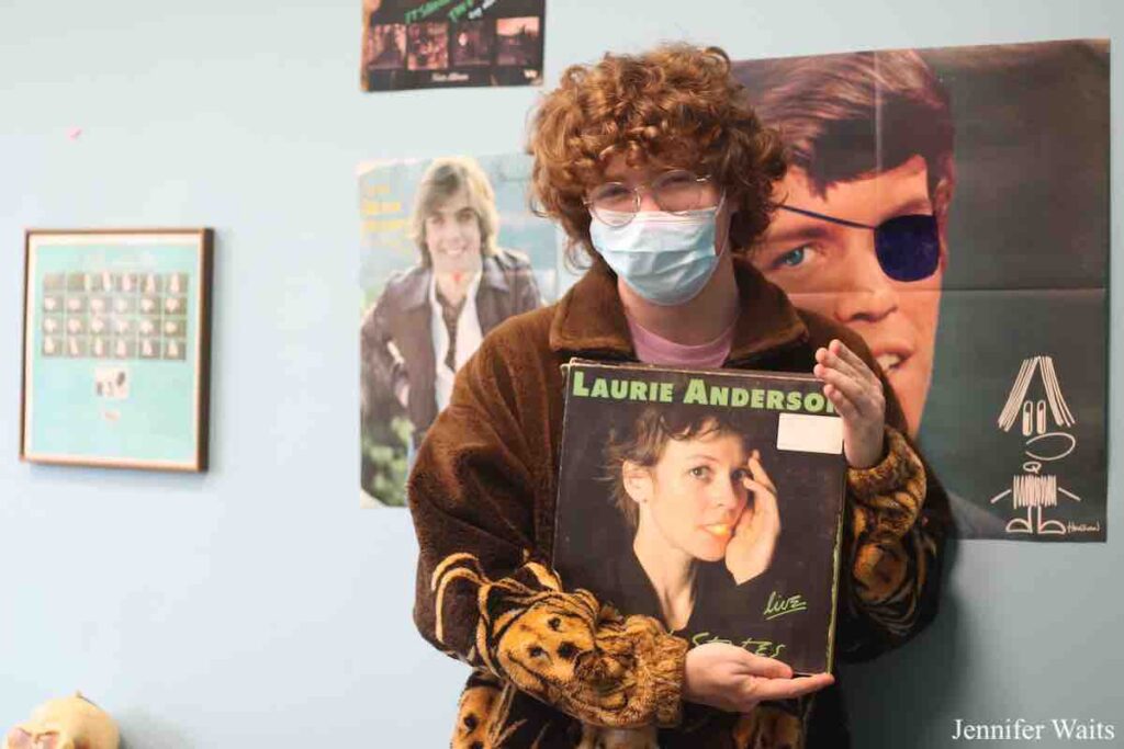 Photo of Henry Burch in college radio station WSLC. He's holding a Laurie Anderson LP. Behind him is a poster of Shaun Cassidy and a poster of a man wearing an eye patch. Photo: J. Waits