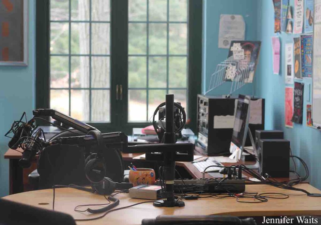 Photo of college radio station WSLC. Pictured is a studio with light blue walls, with flyers posted on them. A desk has microphones attached, with headphones on them. Other audio equipment is on the desk, including a stack of audio players. Photo: J. Waits