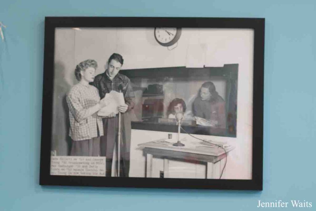 Photo of black and white photo on the wall of college radio station WSLC. Photo on the wall is from the 1940s and is of radio station WSLC. It depicts a man and woman with a script and standing by a microphone. Behind them is a window and we can see two women in that room. 2023 photo by J. Waits