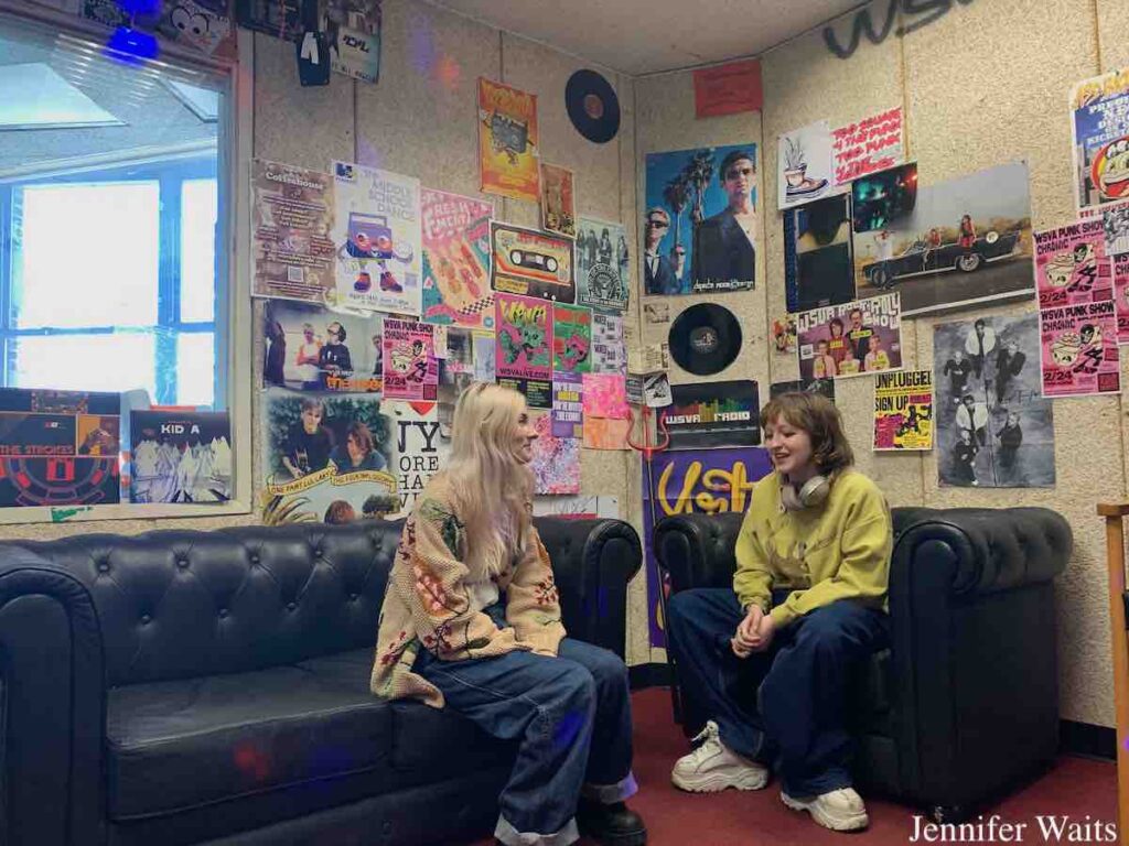 WSVA managers Michelle Mullin and Alice Katok in the studio at the School of Visuals Arts' college radio station in March, 2023. Mullin is on a black couch, Katok sits on black chair. Behind them are poster and flyer-covered walls. Photo: J. Waits