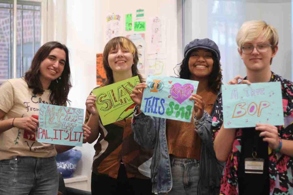 WPIR Pratt Radio Board Members Arzu Oran, Eve Mikkelson, Lili Leoung Tat and Colin Coffey. They are all holding up signs with messages for DJs. They are in the college radio station.
Photo: J. Waits